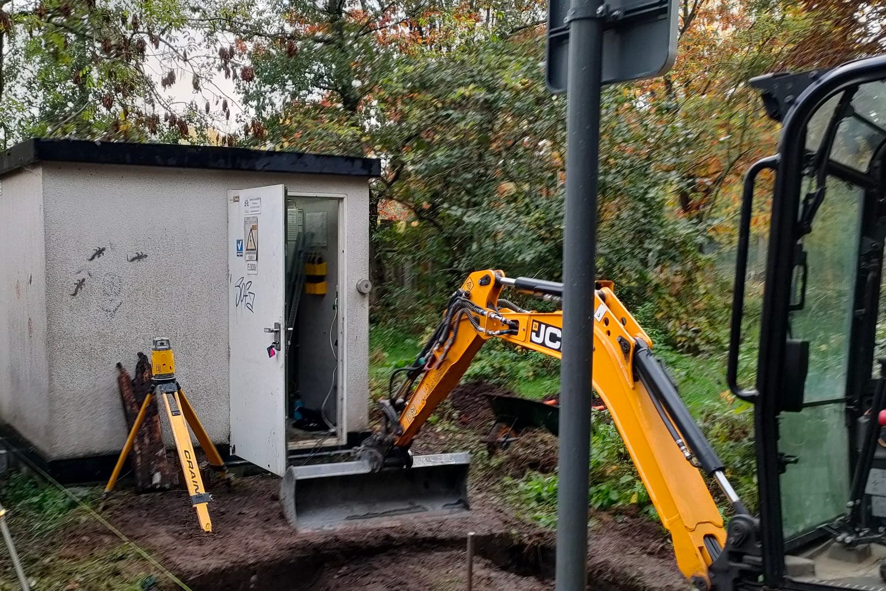 Een distributiecabine met de deur open, met daarnaast een graafmachine die een sleuf trekt voor de nieuwe kabel