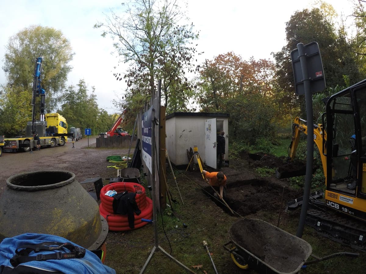 Een werf met een kraanwagen, een graafmachine en kabelhaspels naast een distributiecabine