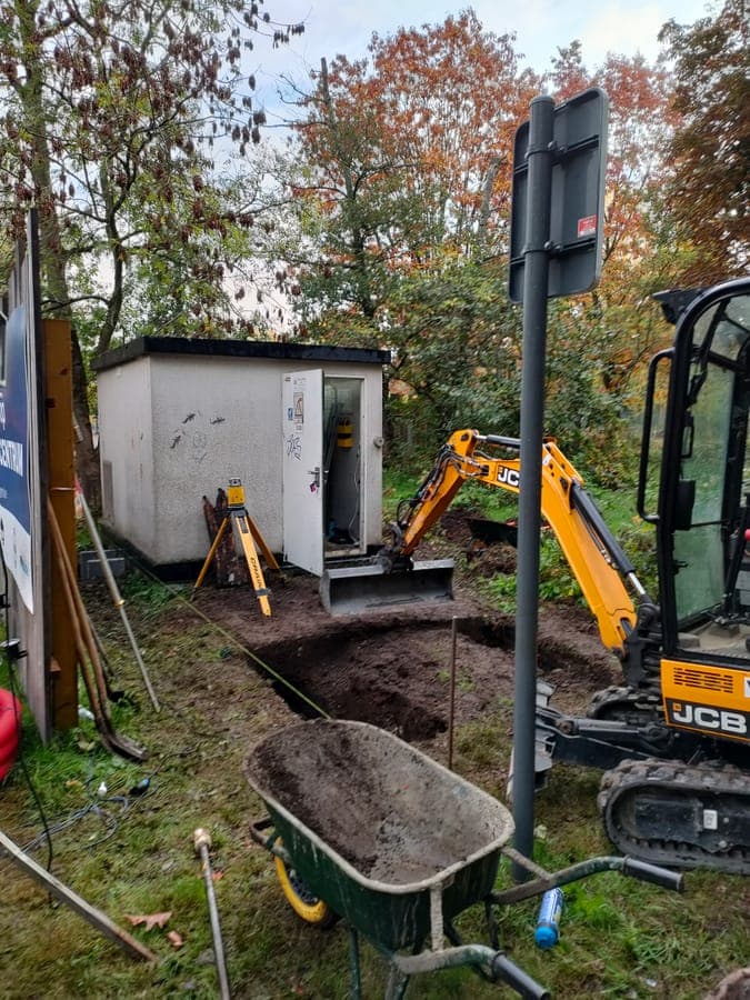 Een graafmachine trekt een sleuf naar een distributiecabine waarvan de deur openstaat
